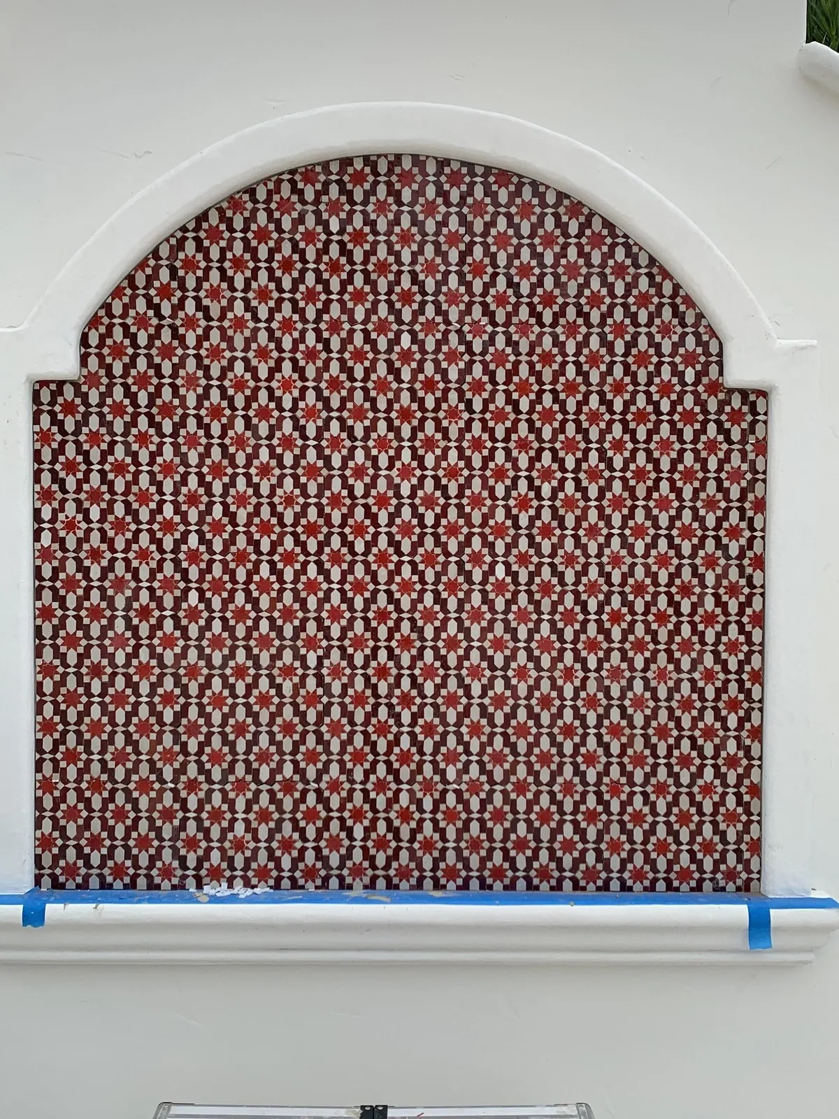 Arched recessed wall panel filled with a repeating red and charcoal star-and-cross patterned tile, photographed during installation with painter’s tape still in place along the base.