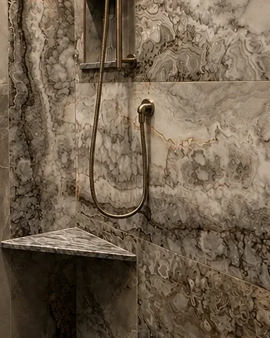 Detail of a triangular corner shower bench with a mitred stone edge, set against continuous veined wall tile beside the valve trim.